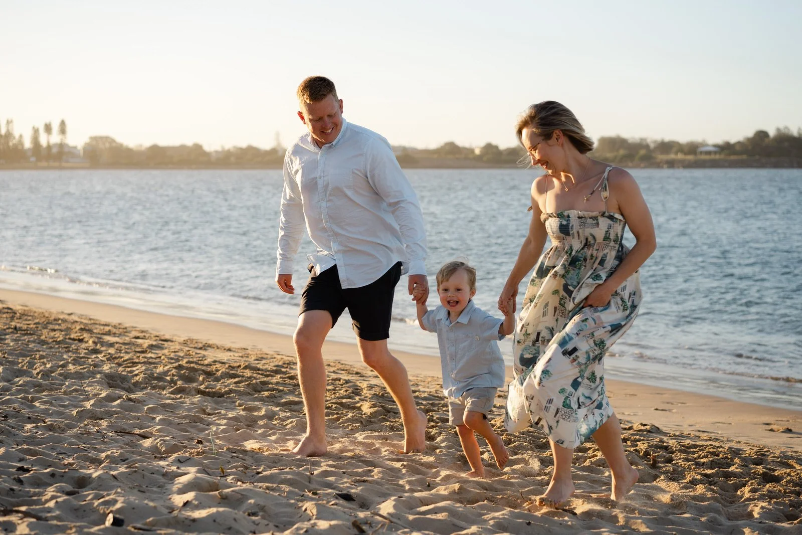 A happy family of three, two adults and a young child, holding hands and walking barefoot on sandy beach near water during sunset.