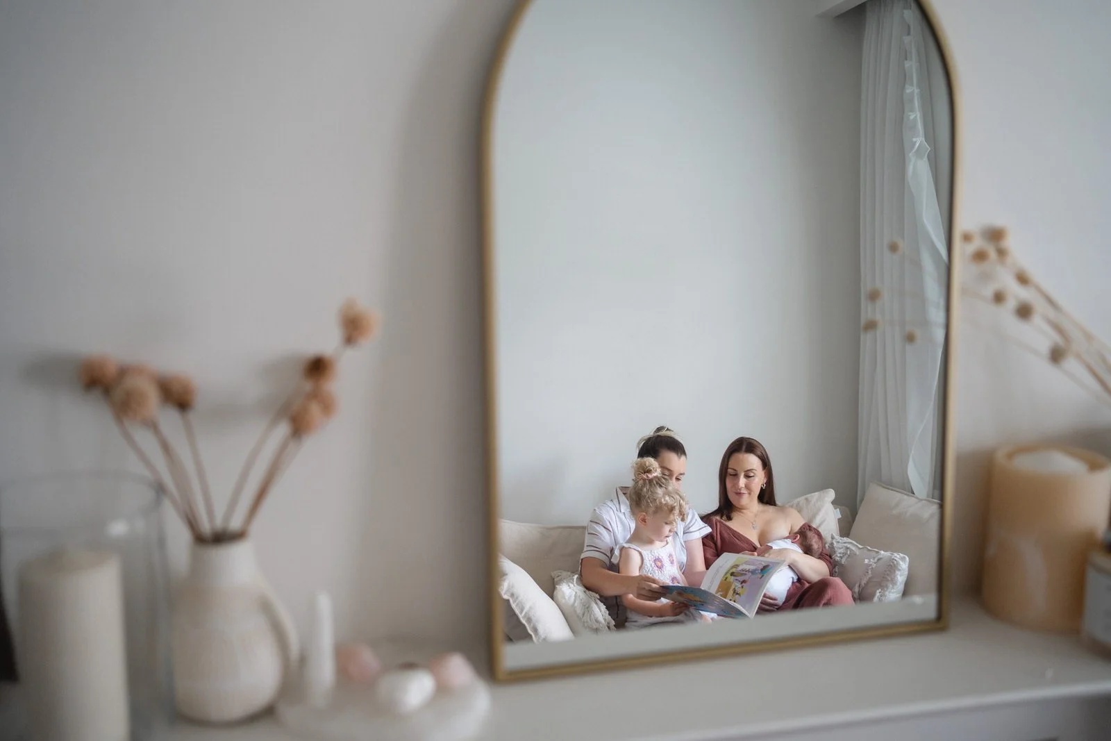 Reflection in a mirror showing three women and two children sitting on a bed, with one woman breastfeeding a baby, as they read a book together.