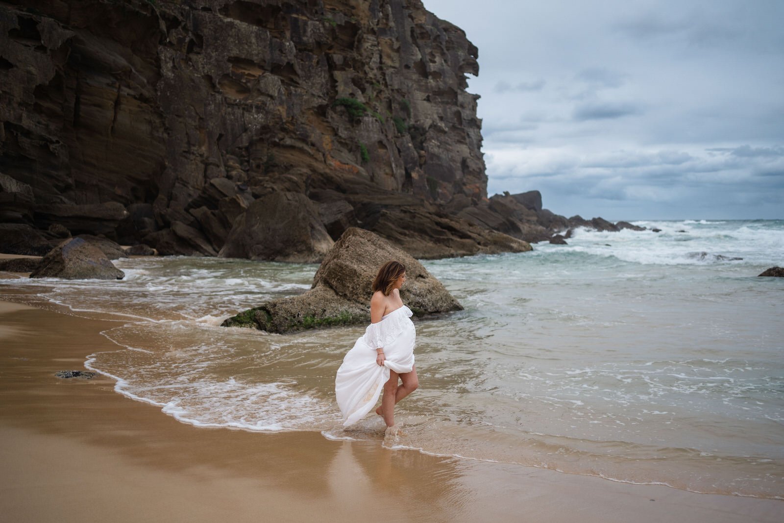 A woman in a white off-the-shoulder dress stands on a sandy beach near the water, with a large rock and dark cliffs in the background under a cloudy sky.