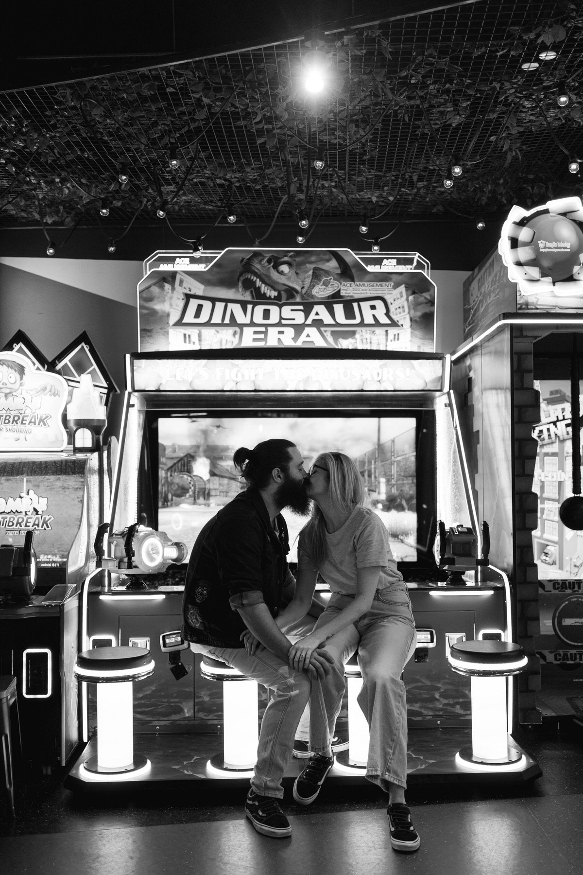 A couple kissing on a bench in front of a dinosaur-themed arcade game in black and white.