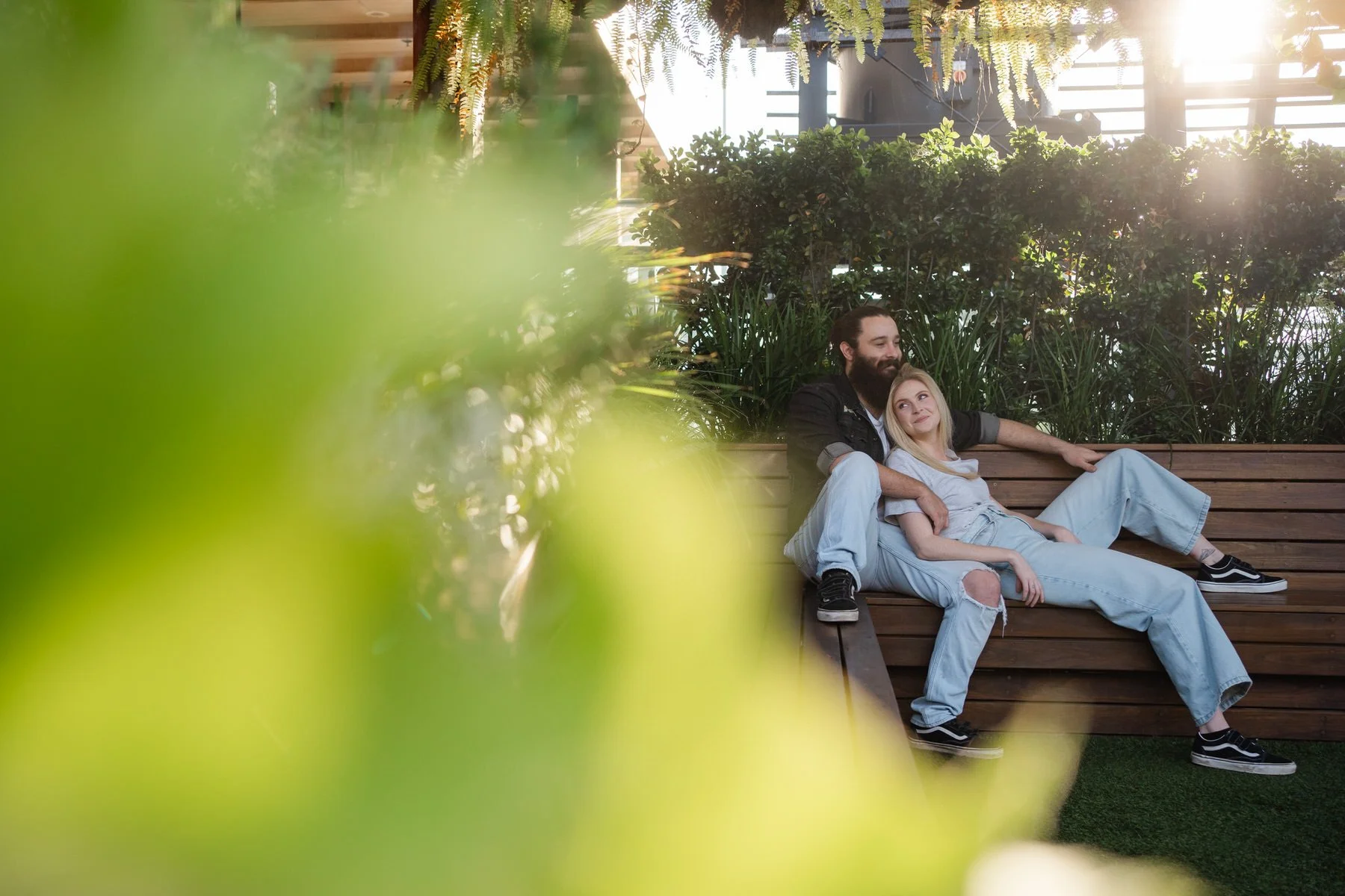 A man with a beard and a woman with blonde hair are sitting on a wooden bench outdoors, surrounded by green plants, with sunlight streaming in from the back.