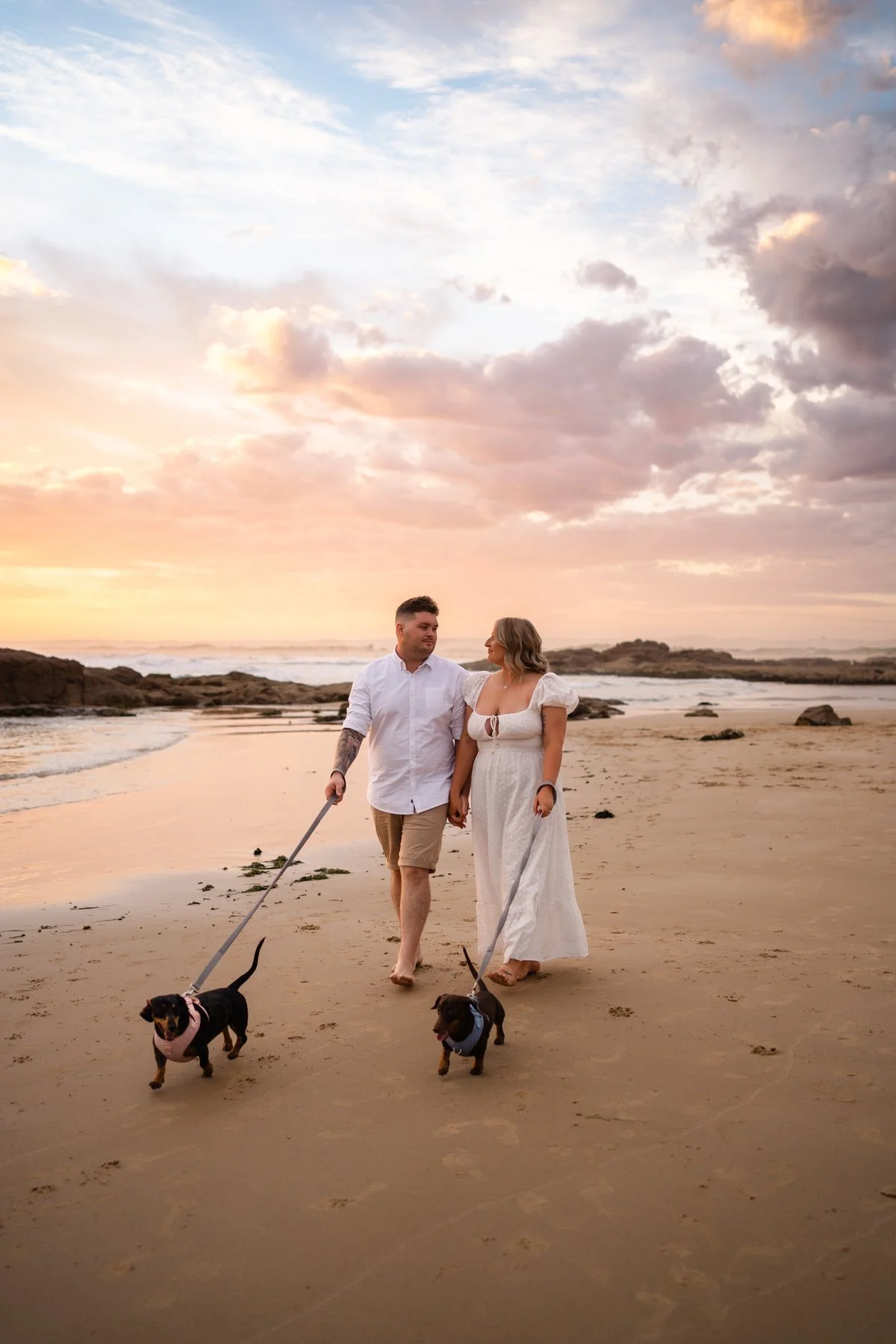 A couple walking on the beach holding hands with two dogs, under a colorful sunset sky.