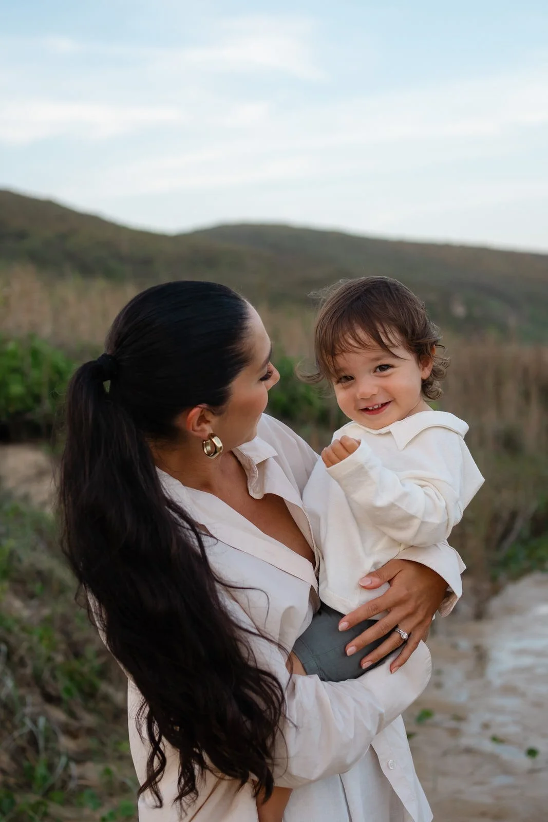 A woman with long dark hair, wearing a white shirt, holding a smiling young boy with light brown hair, also in a white shirt, outdoors near water and grassy hills.
