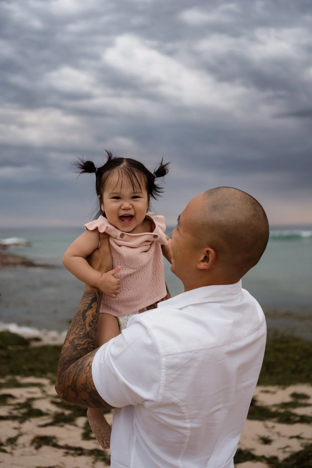 A man with tattoos holding up a smiling young girl on a beach with cloudy skies.