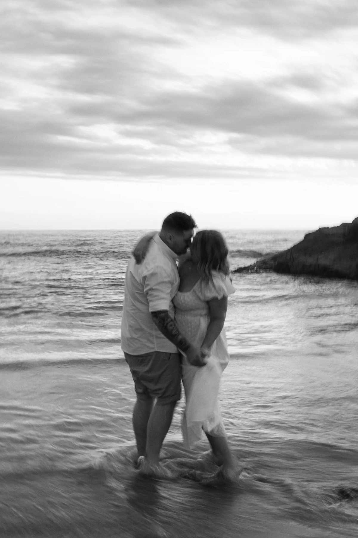 A couple standing in the shallow water at the beach, kissing, with the ocean and cloudy sky in the background.