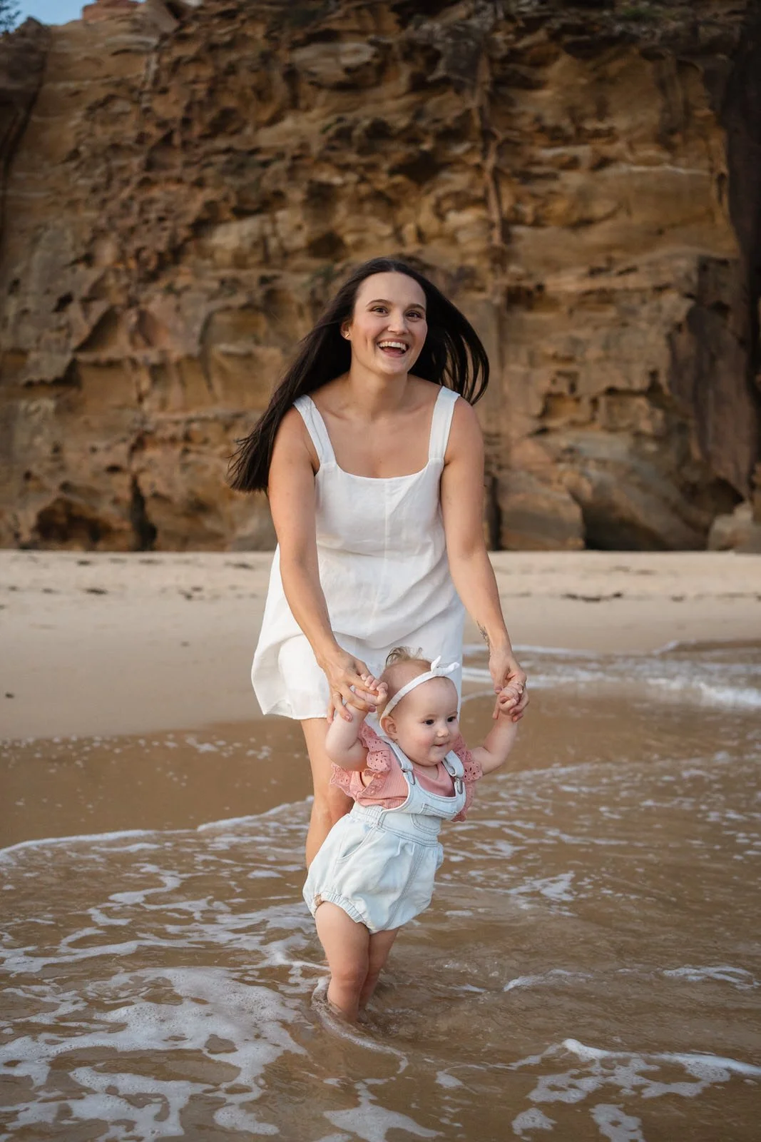 A woman and a young girl standing in the shallow surf at a beach, with rocky cliffs in the background, smiling and holding hands.