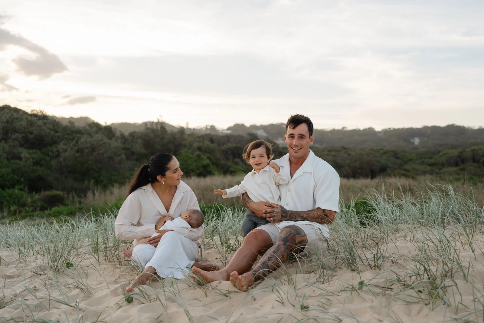 Family of four sitting on sandy beach with grass, ocean and green hills in the background. The mother is breastfeeding a baby, the father is holding a toddler, all wearing light-colored clothing.