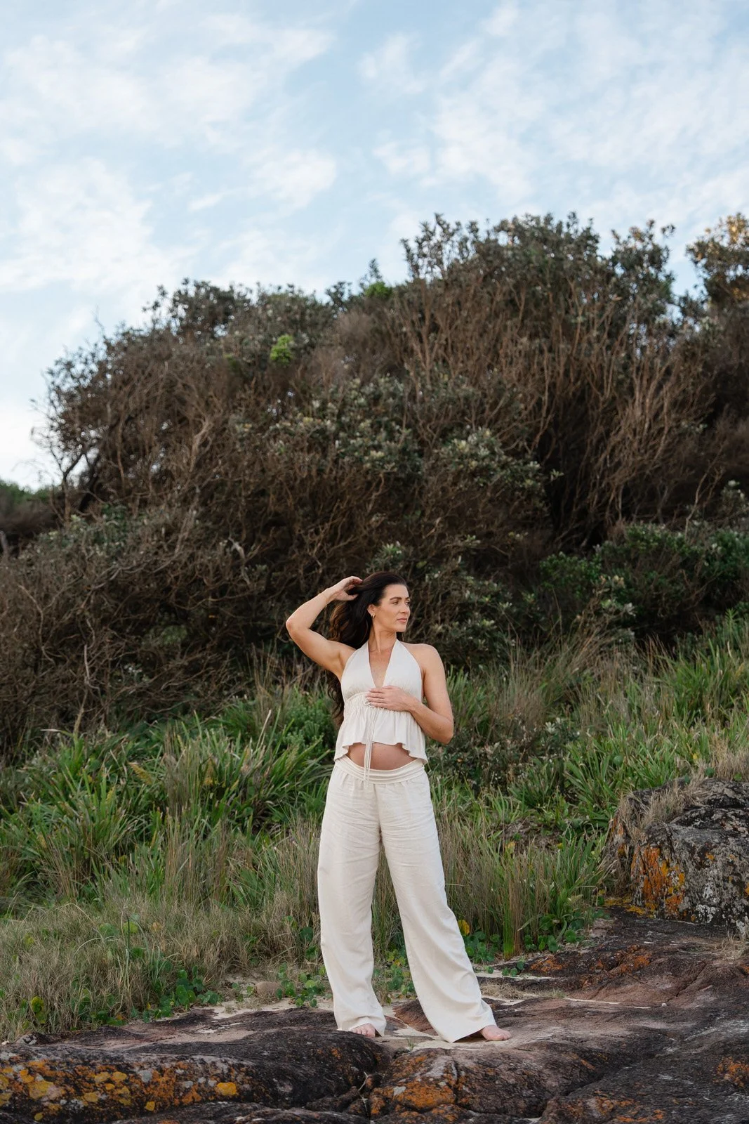 Woman in a cream-colored halter top and wide-leg pants standing on rocks near greenery and dried bushes, with a partly cloudy sky above.