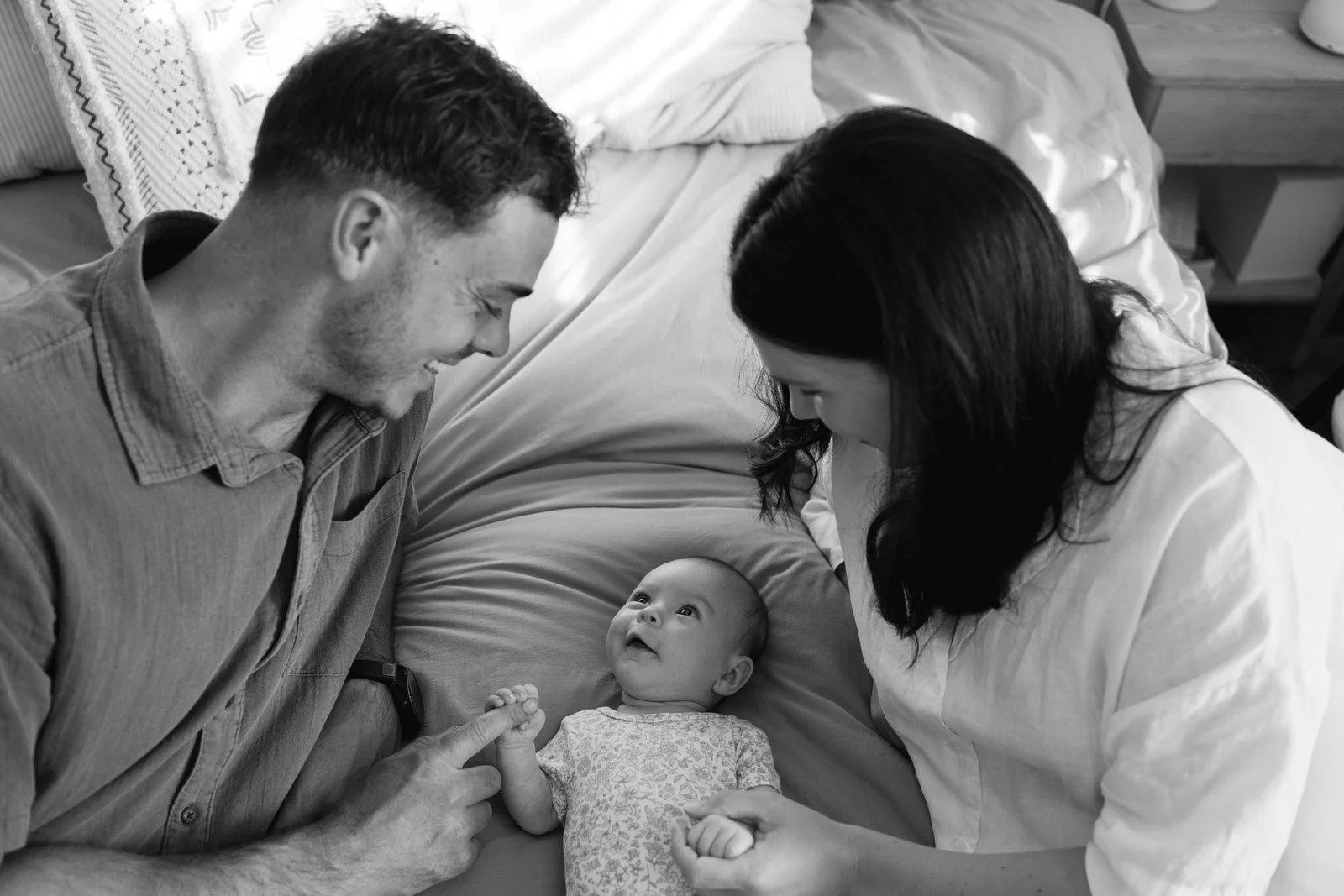 mother and father laying on a bed on either side of their baby girl, holding her hands and smiling at her