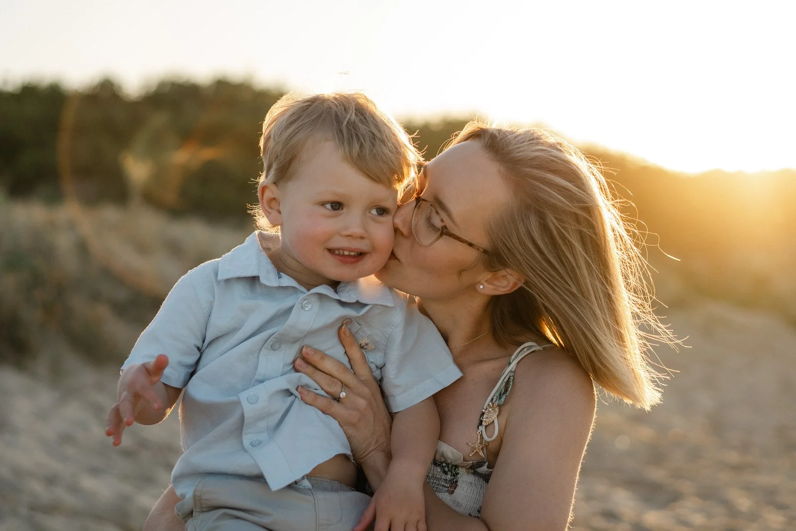 A woman kissing a young boy on the cheek outdoors during sunset, with sandy terrain and vegetation in the background.