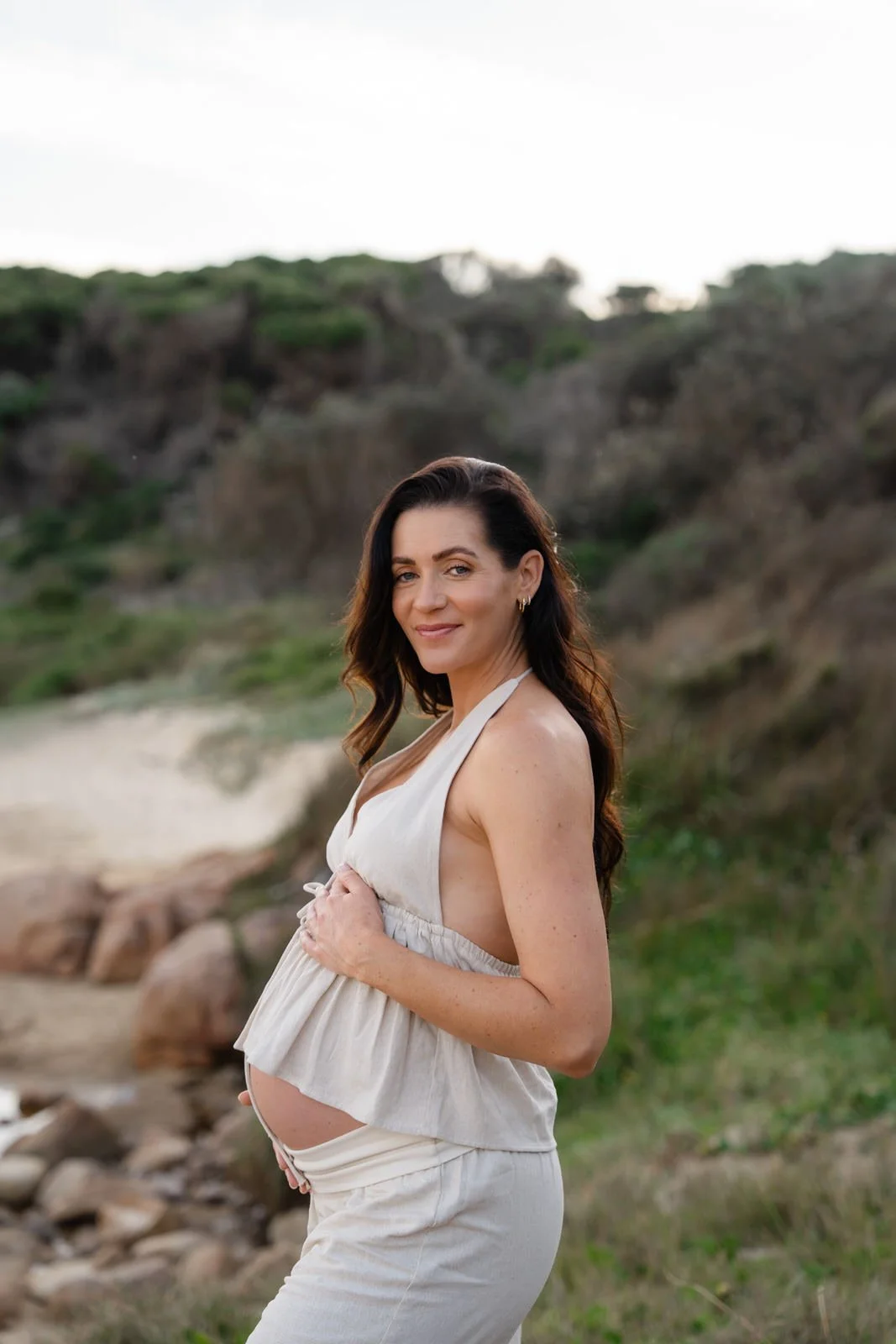 Pregnant woman standing on the rocks at the beach, holding her belly and smiling