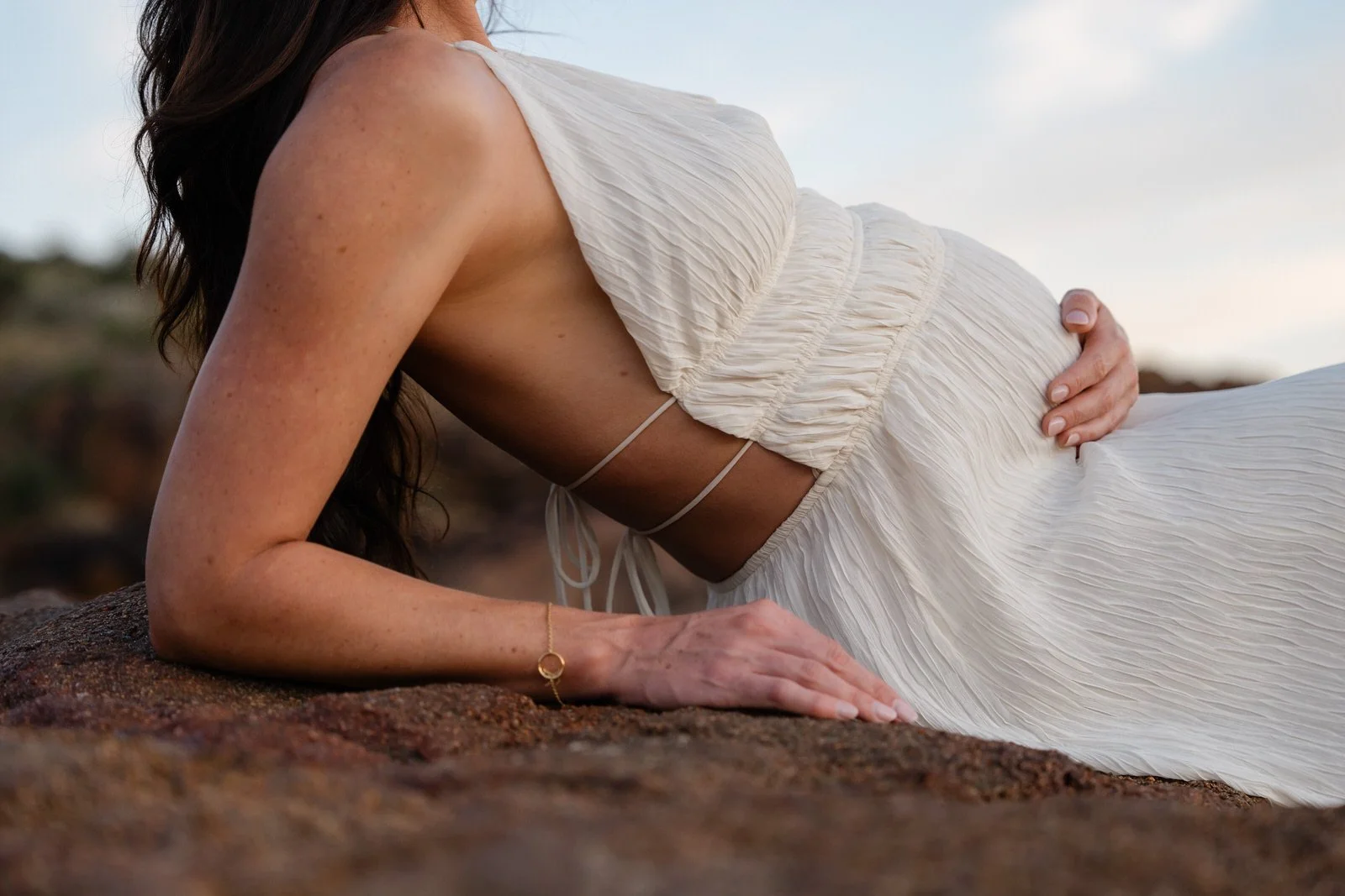 A woman in a white, flowing dress pushing herself up on her hands and knees on a rocky surface outdoors.