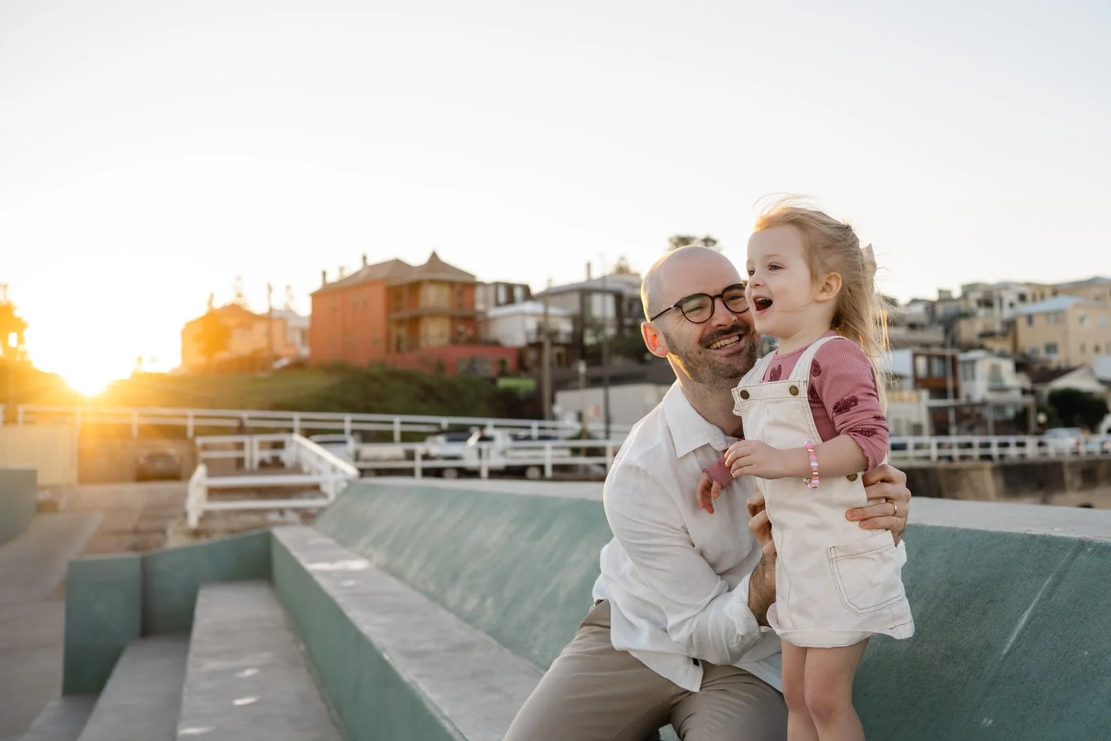 A man with glasses holding a young girl with blonde hair, both smiling and enjoying the sunset outdoors in a coastal area with houses and a walkway in the background.