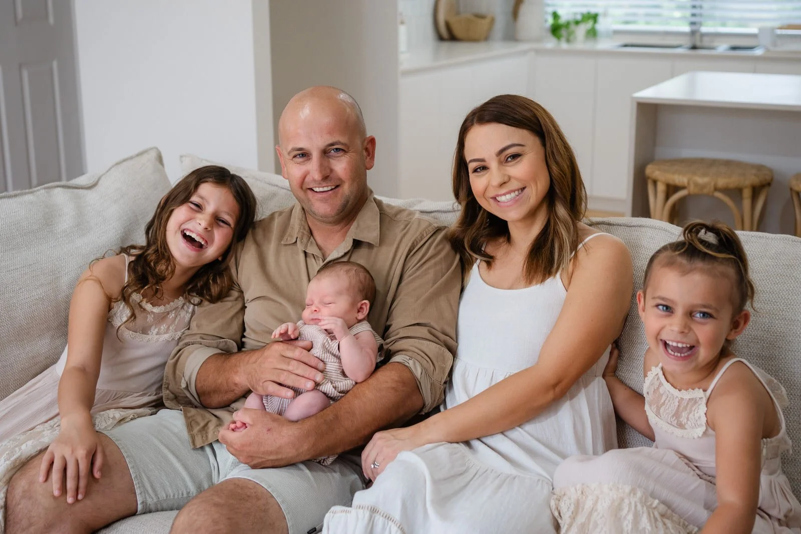 A happy family of five sitting on a beige couch in a bright living room. The family includes a man holding a newborn, a woman, and three young girls, all smiling.