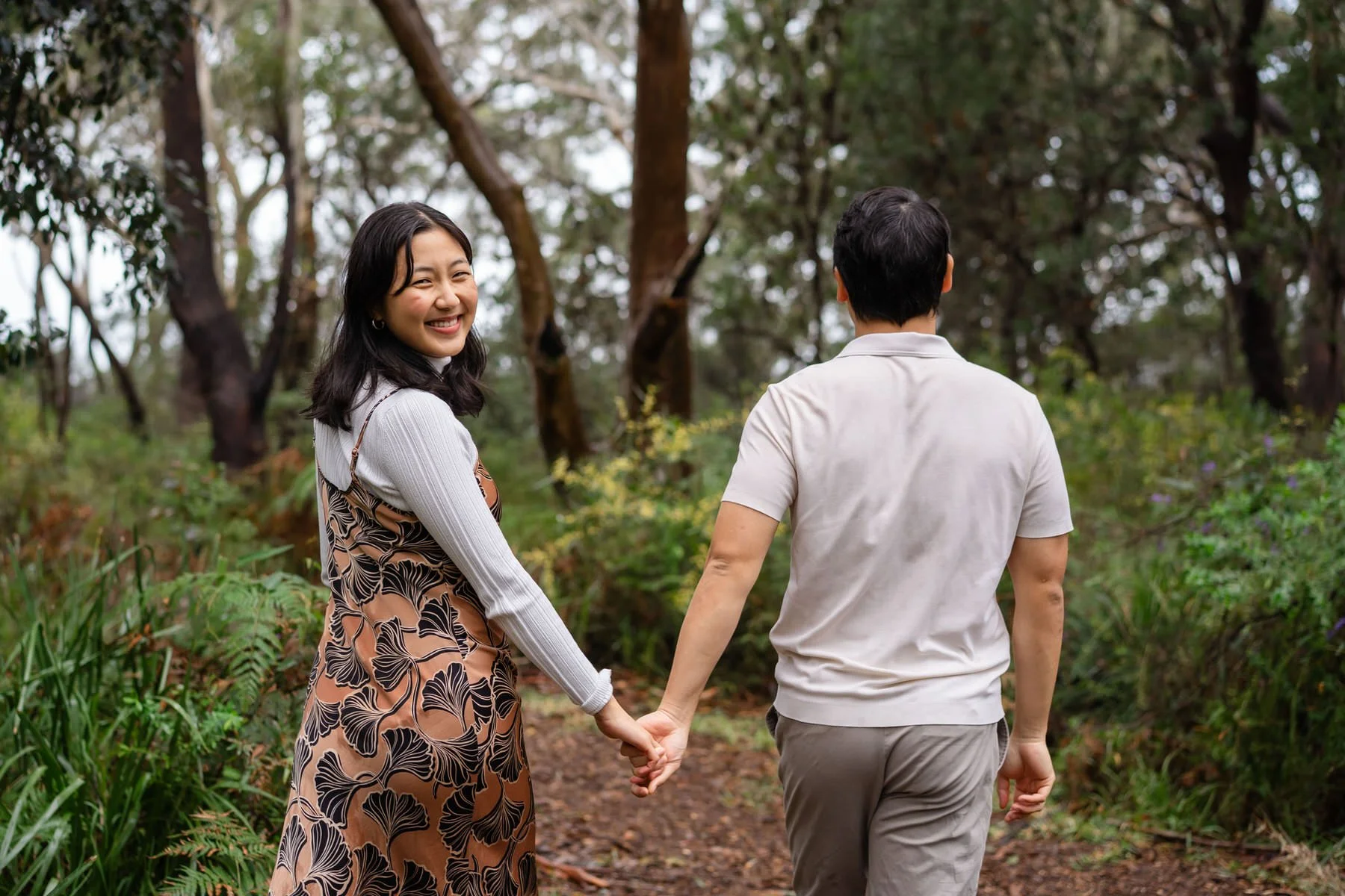 A woman with long, dark hair smiling and holding hands with a man as they walk through a forested trail for their engagement photoshoot in Port Stephens.