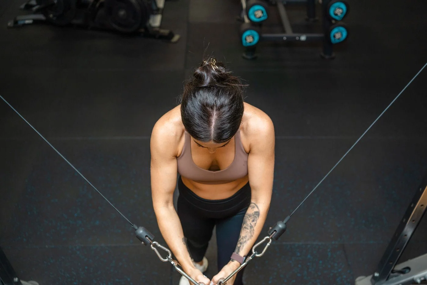 Woman wearing a sports crop top, holding onto the cable machine with both arms and looking down
