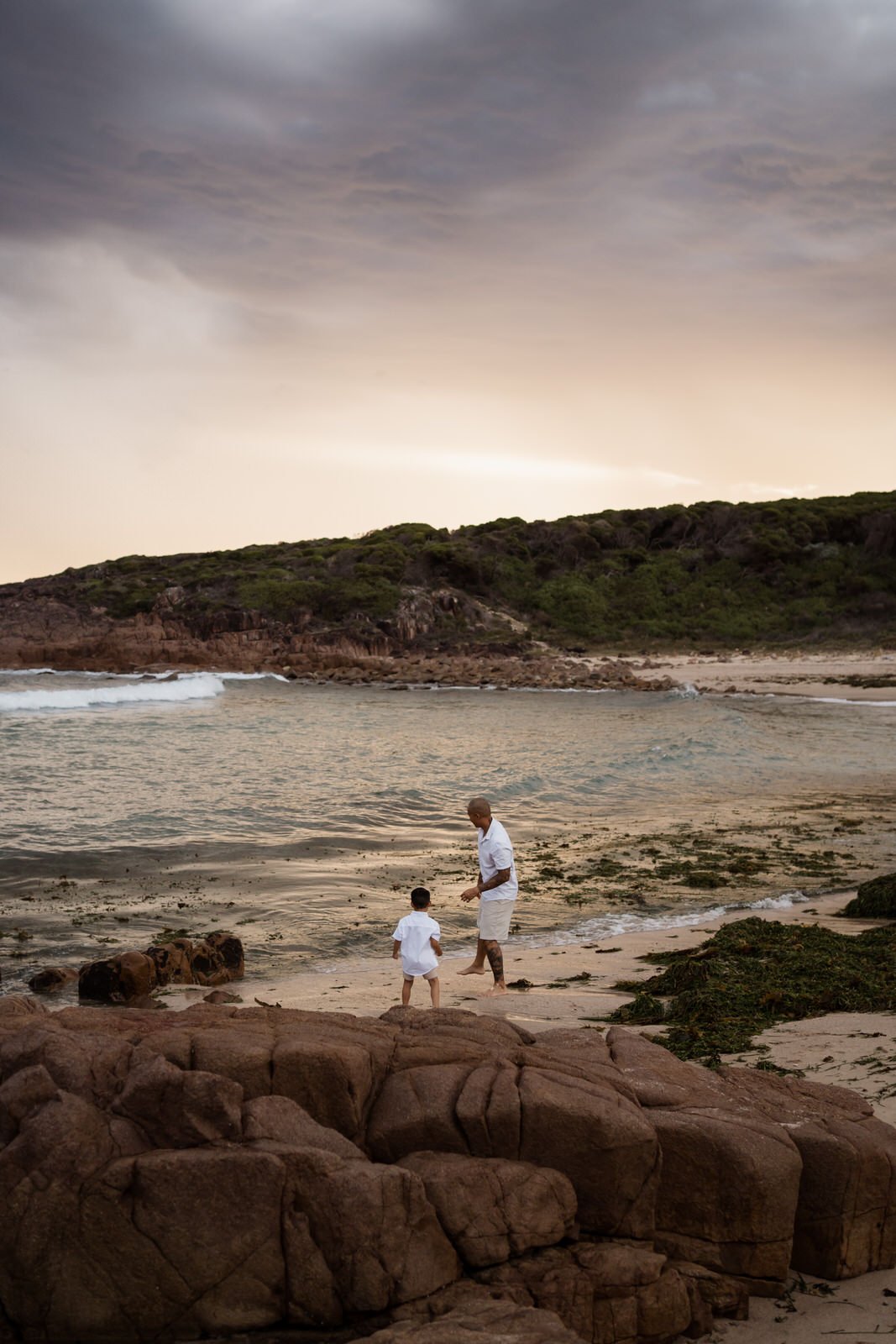 A man and a young child walking along the shoreline of a beach with rocks in the foreground and green hills in the background during sunset or dusk.