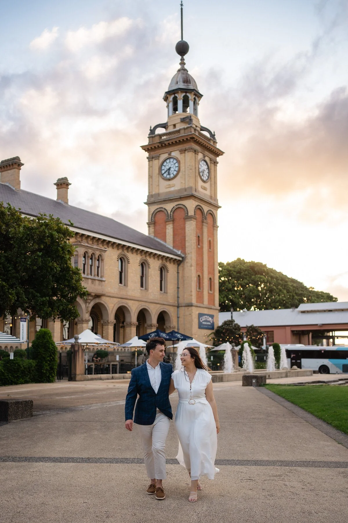 A couple dressed in formal attire walking arm in arm in front of a historic clock tower at sunset.