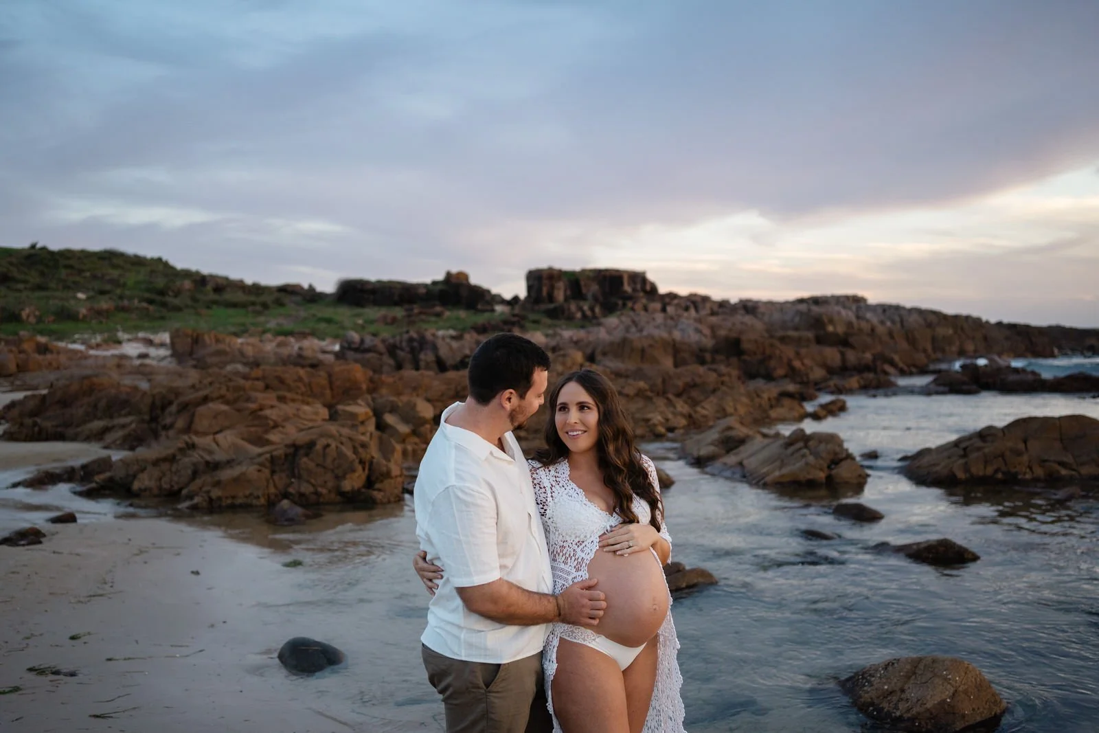 A pregnant woman and a man standing on the beach near rocks, with rocky hills in the background during sunset.