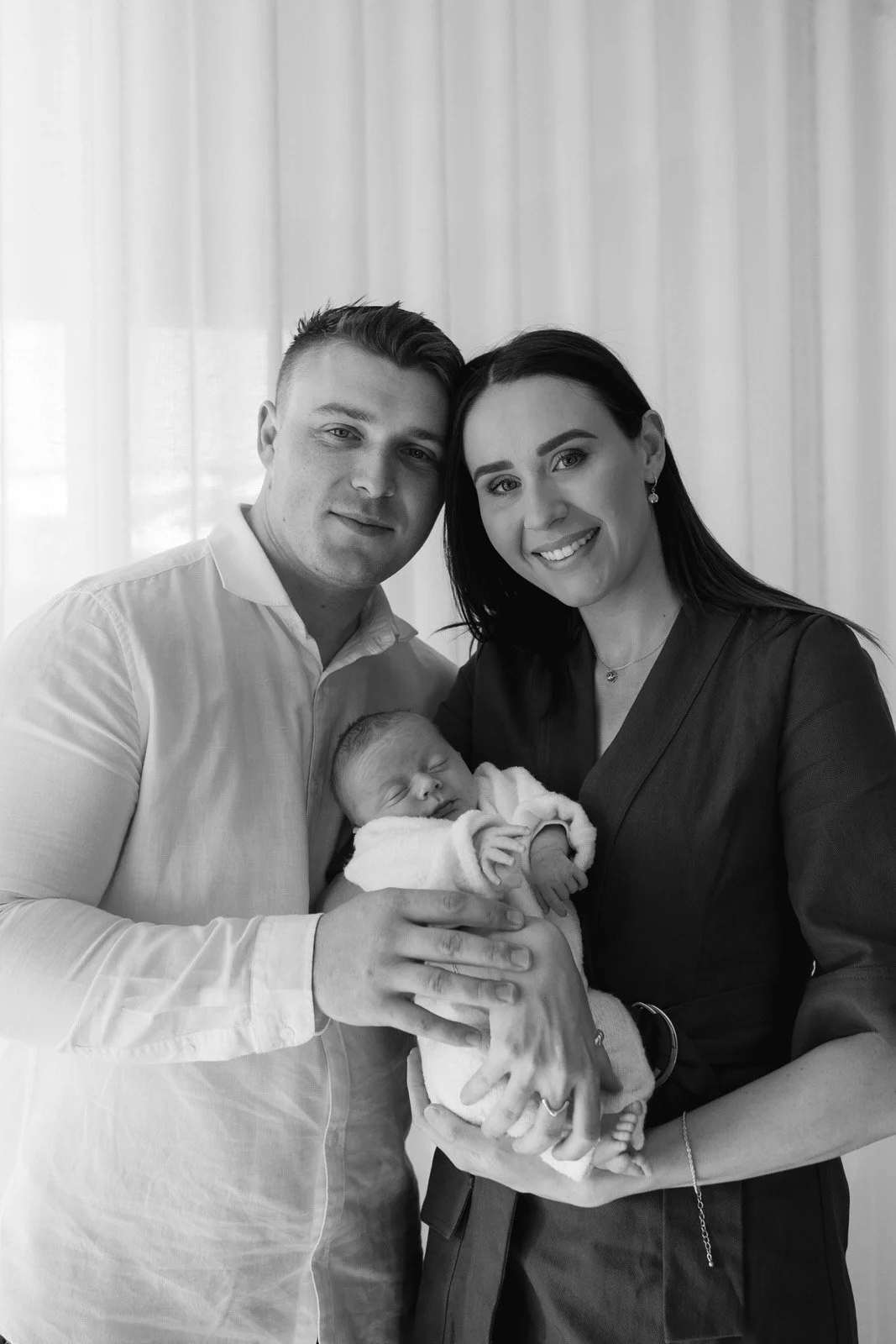 Black and white photo of a smiling woman and man holding a sleeping newborn baby, indoors with curtains in the background.