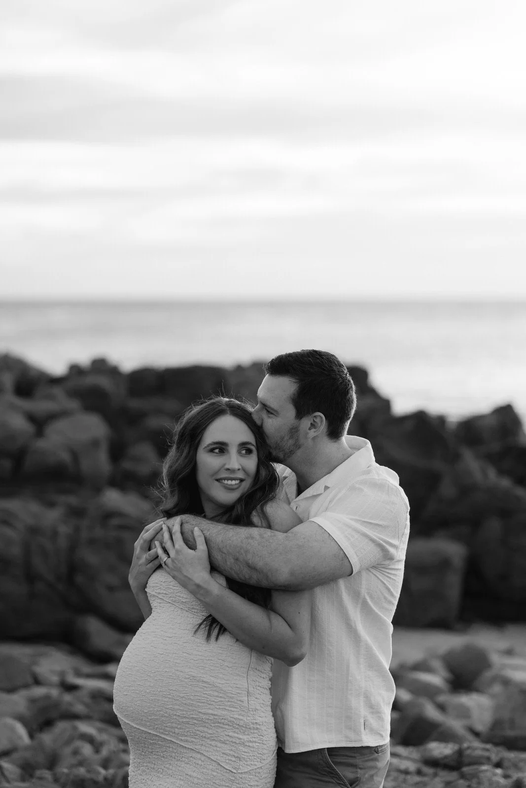 A black-and-white photo of a couple on a rocky beach, with the man kissing the woman's temple and both hugging, near the shoreline with water and cloudy sky in the background.