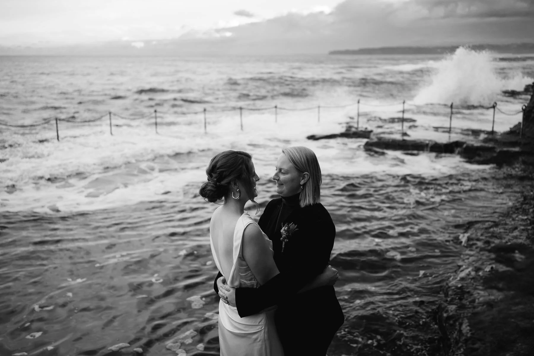 two brides embracing each other, while standing near the bogey hole in Newcastle NSW
