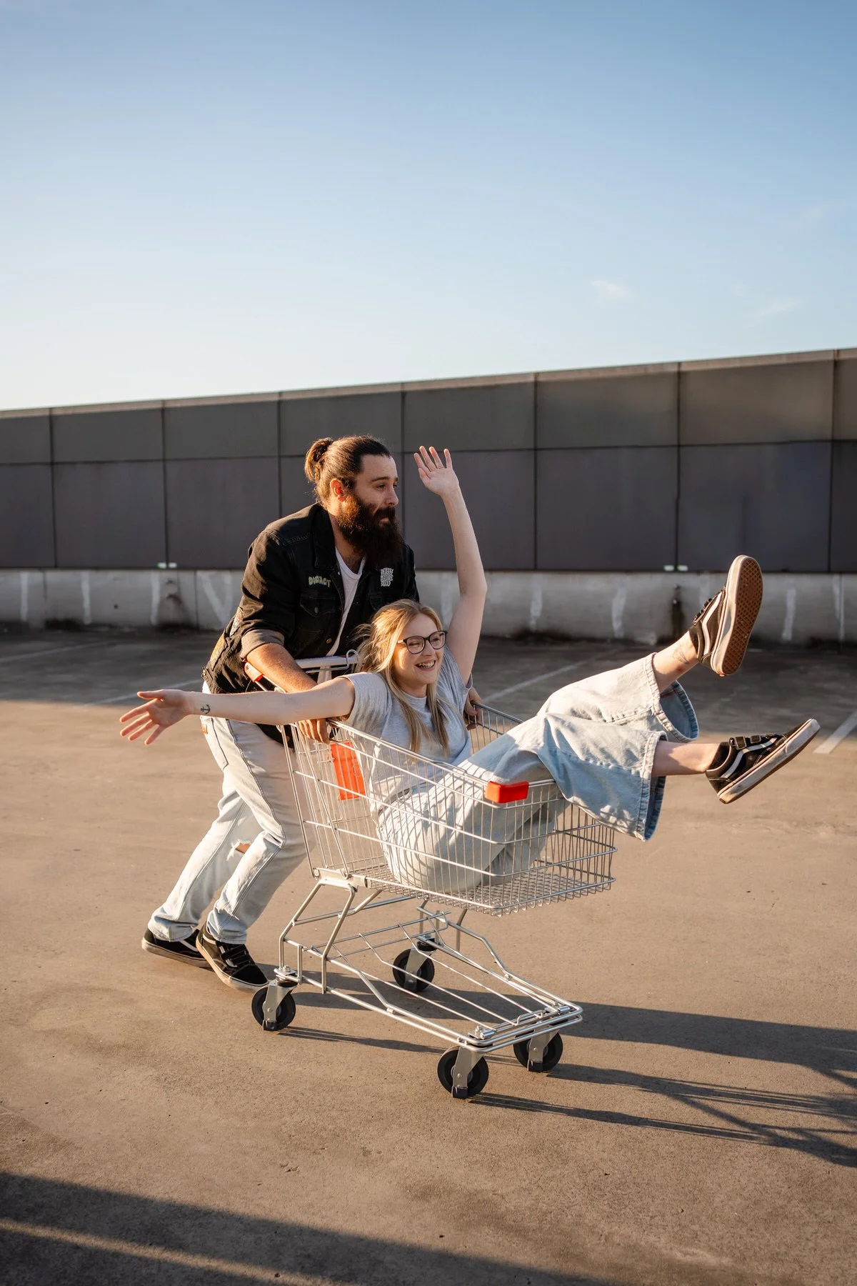 A man pushing a shopping cart with a smiling girl inside, her arms stretched out wide and one arm raised, on a rooftop parking lot during sunset.