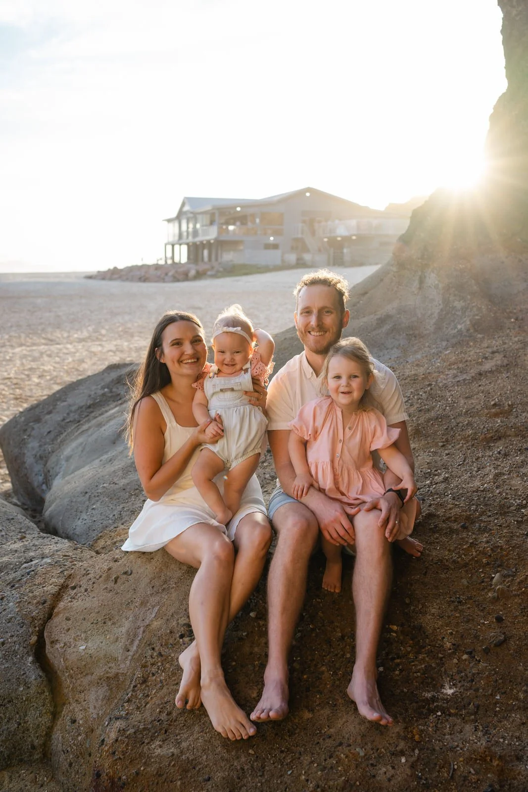 A happy family sitting on rocks at the beach during sunset, with a house in the background.