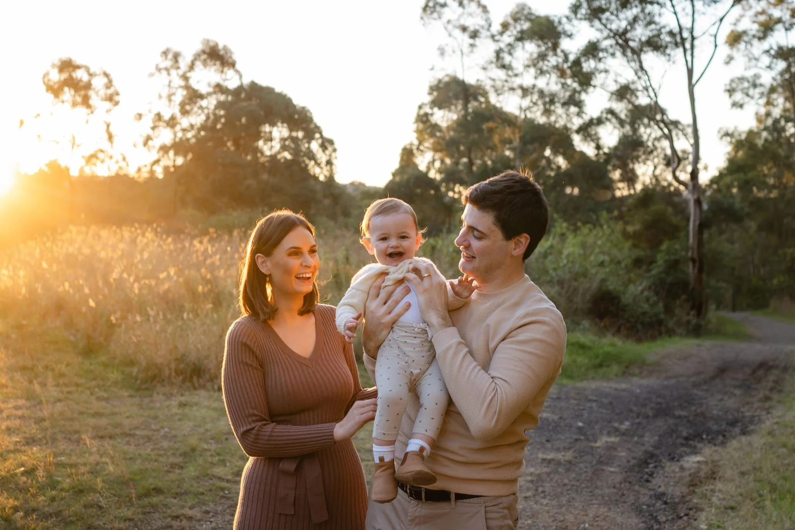 Family of three enjoying sunset outdoors, with a woman, a man holding a smiling baby, trees in background, warm lighting.