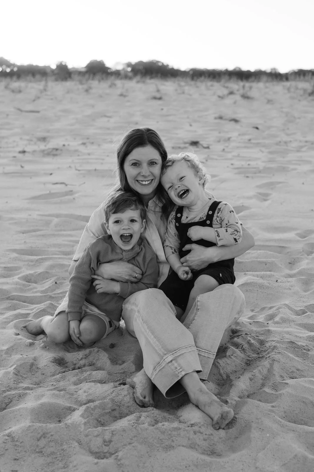 A woman sitting on the sand with two young children, all smiling and laughing together on a beach, black and white photo.