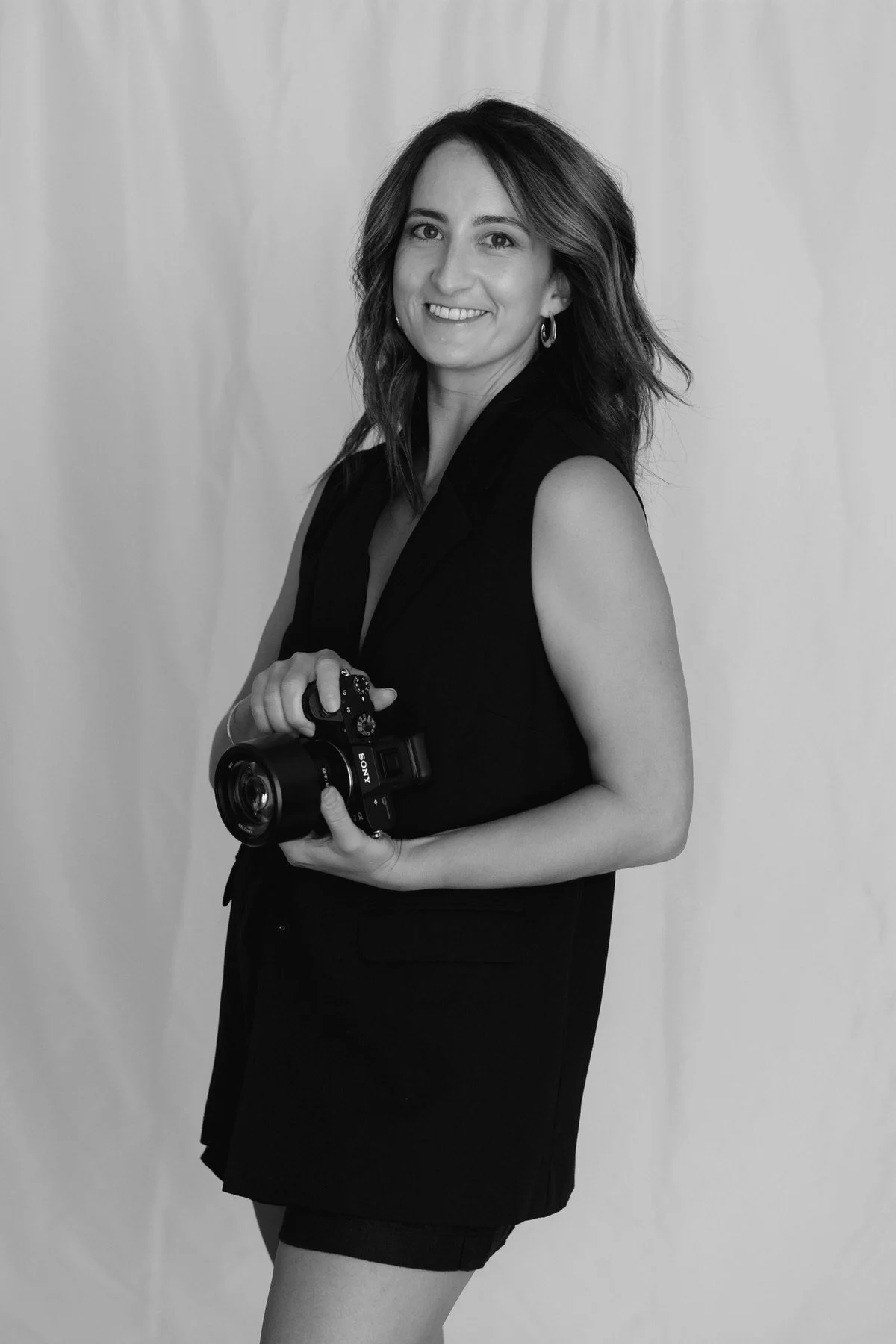 Black and white image of a woman wearing a black vest, holding a camera and smiling