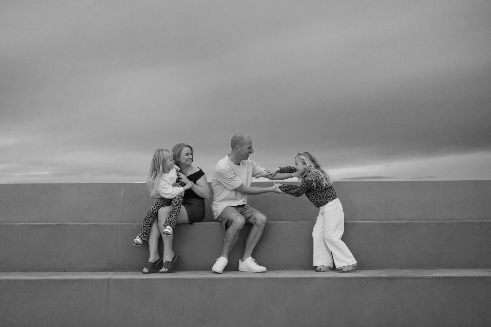Black and white photo of a family playing tug-of-war with a toy on a concrete ledge outdoors, with a cloudy sky in the background.
