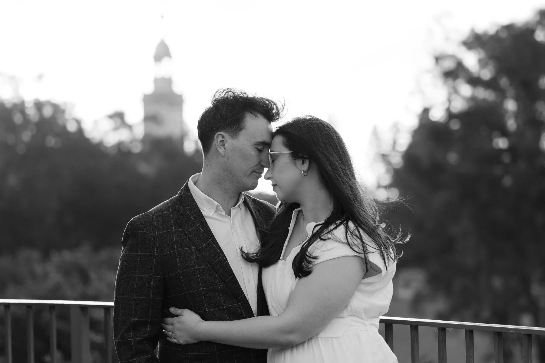 A black and white photo of a man and woman standing close together with their foreheads touching, on a bridge outdoors with trees and a distant building in the background.
