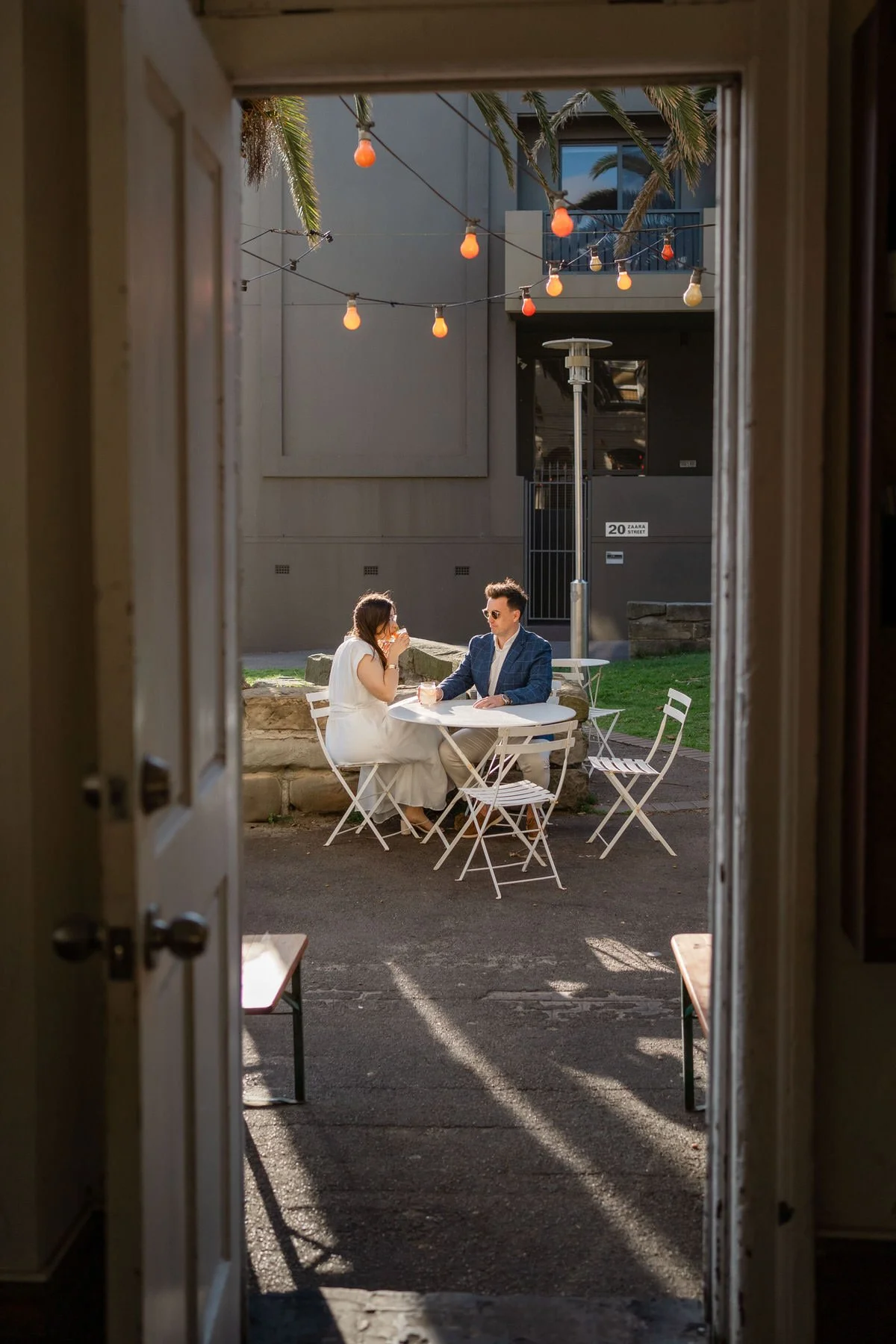 A couple is sitting at a white outdoor table, having drinks, seen from inside a building through a doorway. The scene is set in the late afternoon or early evening with string lights overhead and palm trees in the background.