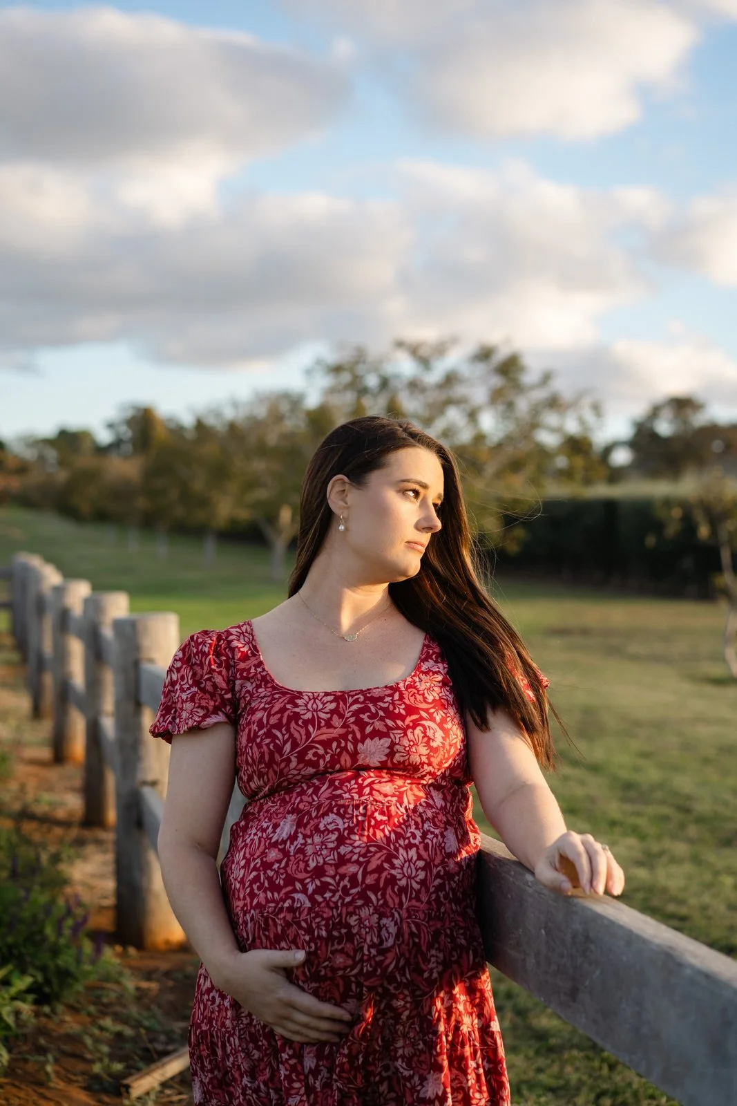 A pregnant woman in a red floral dress stands outdoors near a wooden fence, holding her belly and looking into the distance with a peaceful expression, during golden hour sunlight.