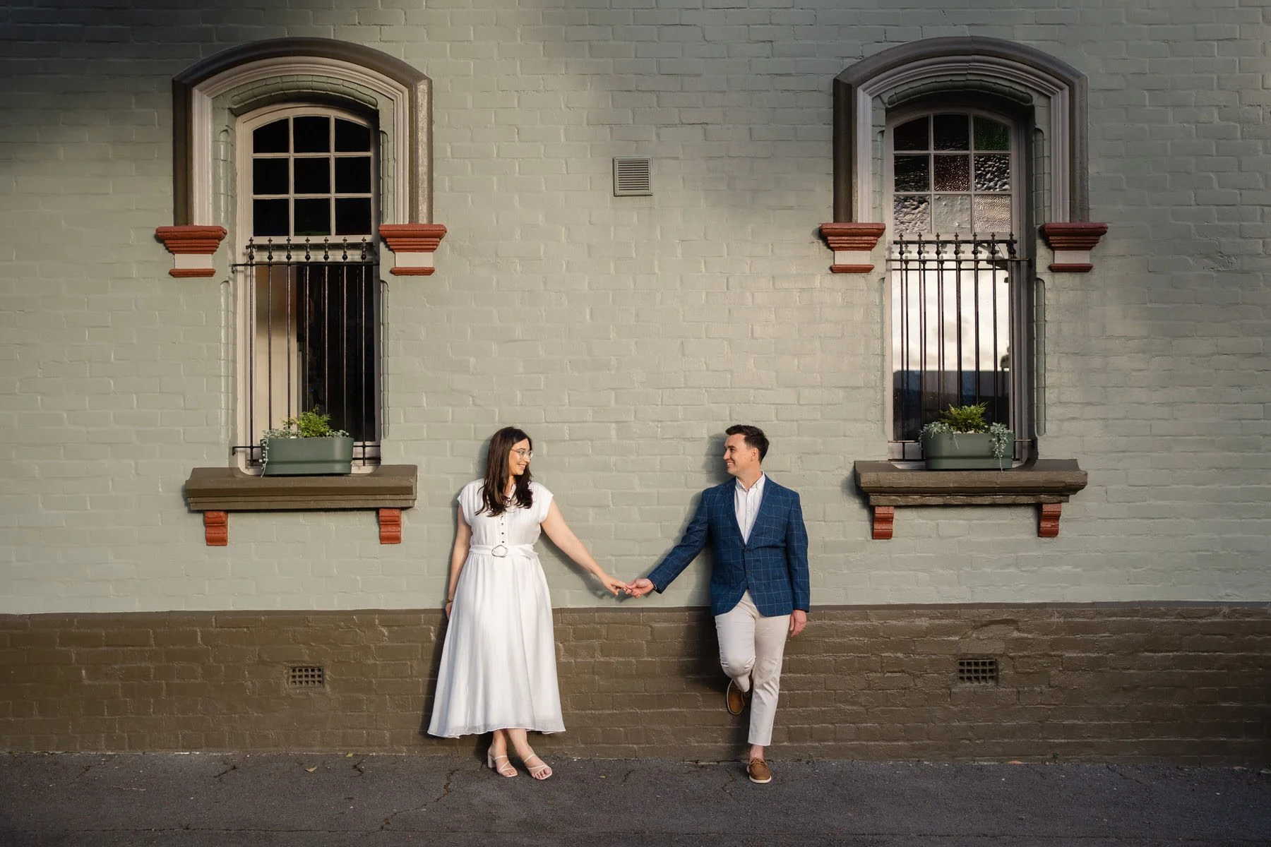 A man and woman holding hands and smiling while leaning against a brick wall with two windows behind them.