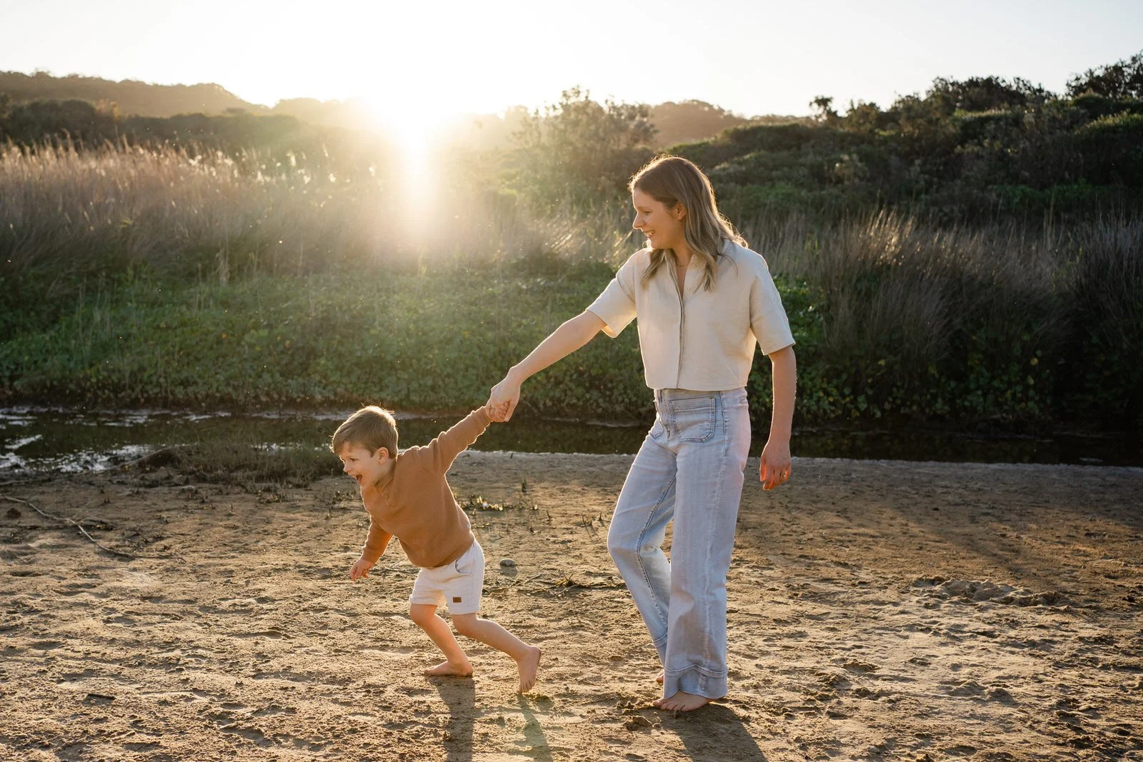 A woman and a young boy playing on a sandy beach near a river at sunset, with the woman holding the boy's hand as he runs.