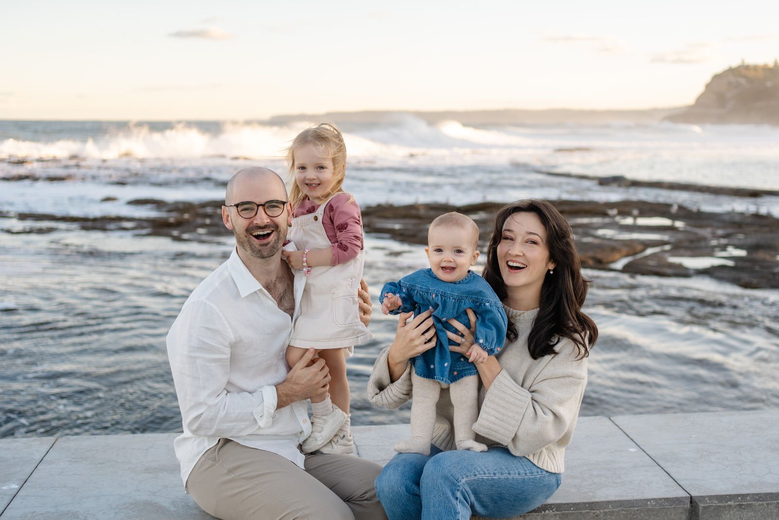 A happy family of four at the beach, with the father holding a girl and the mother holding a baby, all smiling at the camera with the ocean and rocky shoreline in the background.