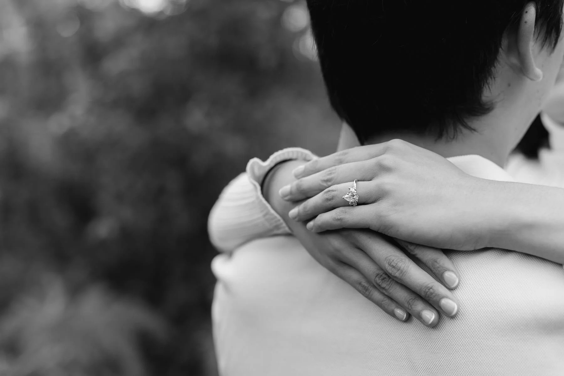 A woman with a ring on her finger is hugging a man from behind, with her hands resting on his shoulder, in a black and white photograph.