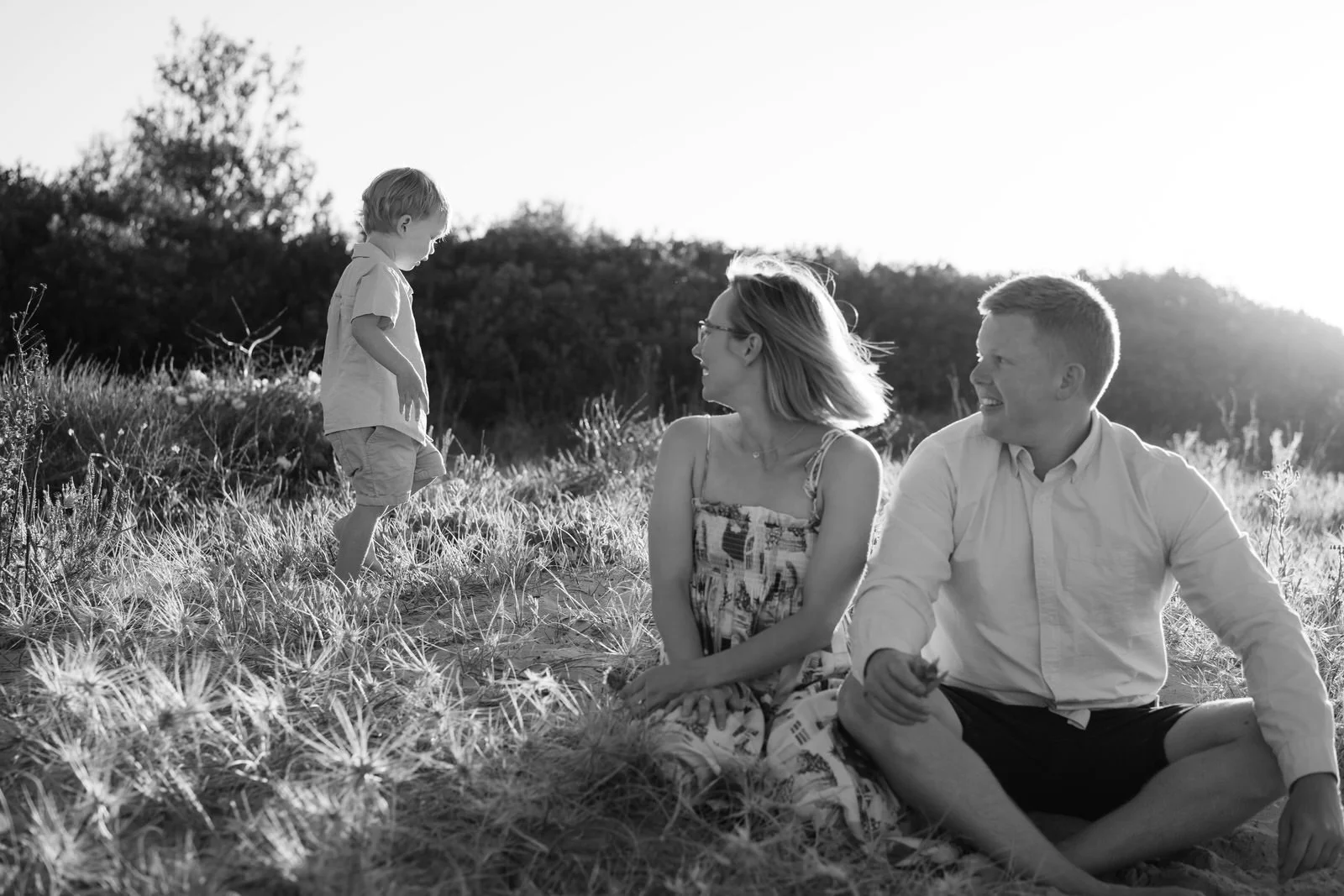 A family of three sitting on the beach, with a young boy approaching them, all smiling and enjoying the moment.