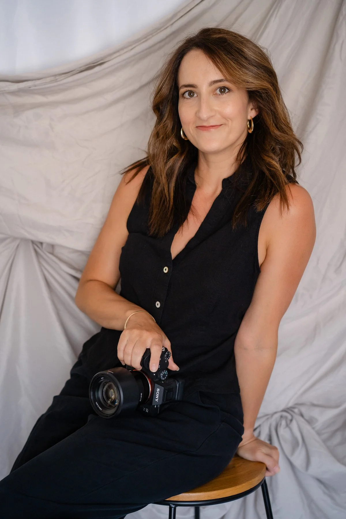 Woman sitting on a stool, wearing all black and holding a camera