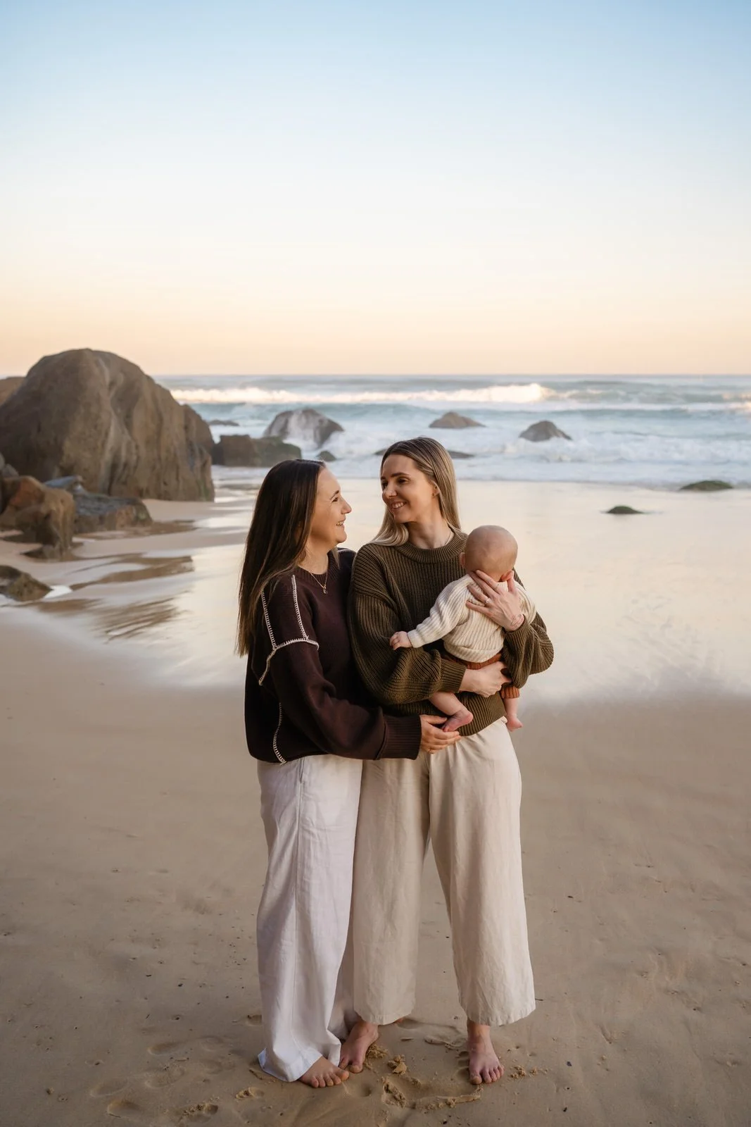 Two women and a baby standing on a sandy beach with rocks and ocean waves in the background during sunset.