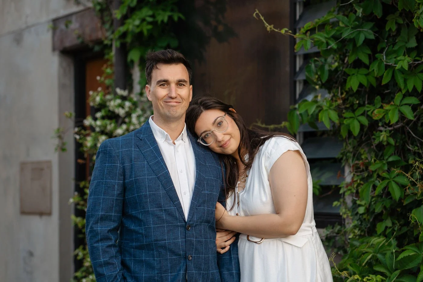 Woman leaning on her husbands shoulder, wearing a white dress and both looking and smiling at the camera