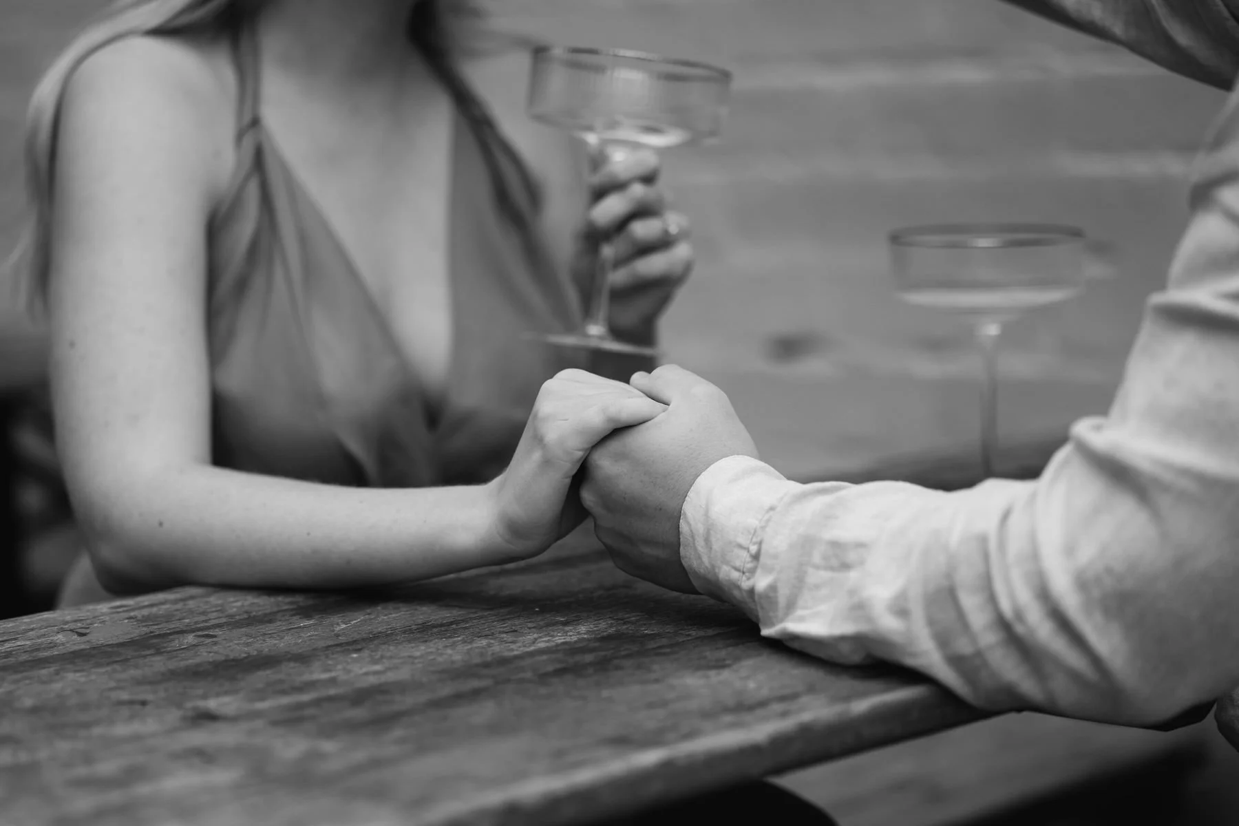 Black and white photo of a woman and a man holding hands on a wooden table, with the woman holding a glass of wine in her other hand.