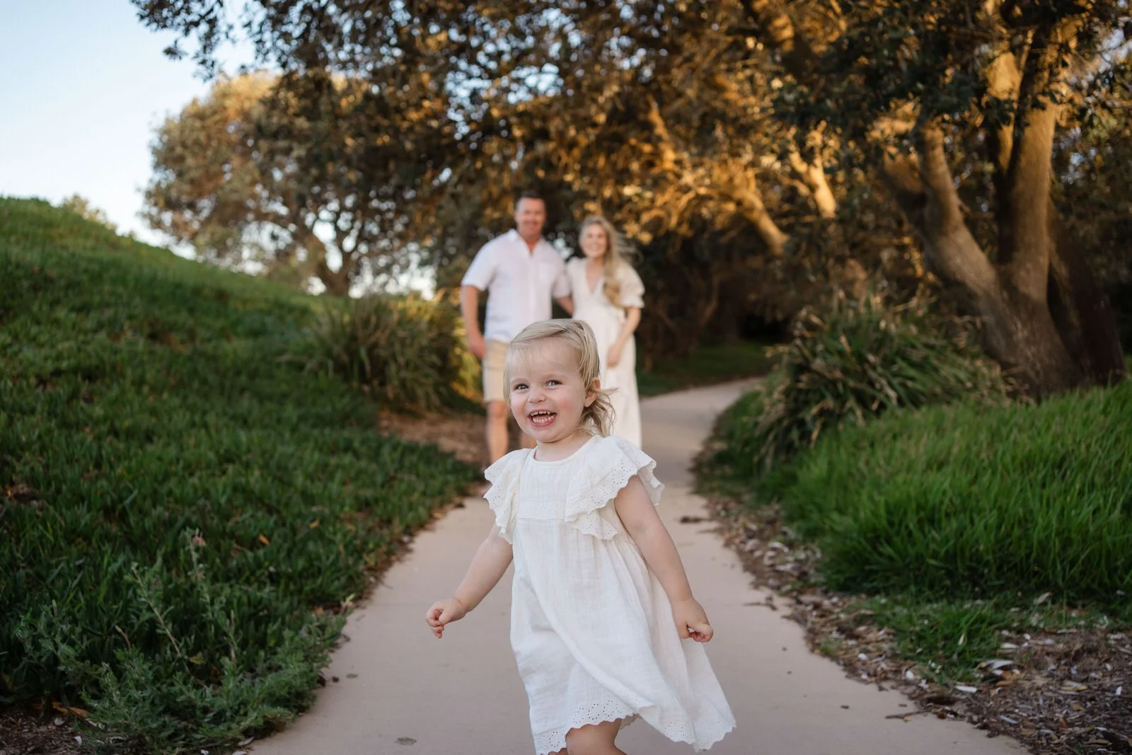 A young girl in a white dress smiling and running on a winding path, with a man and woman in casual attire walking behind her in a park setting during late afternoon at Birubi Beach