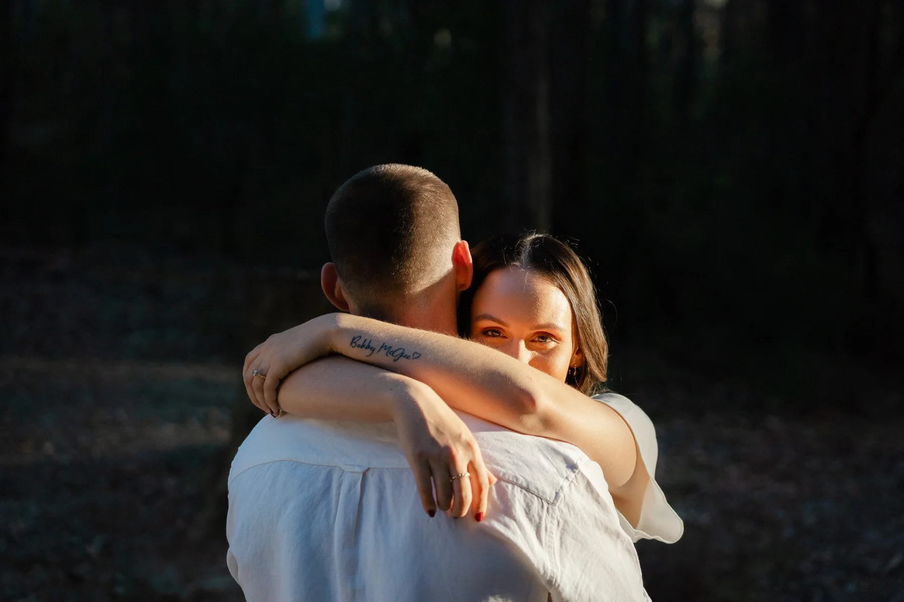 A woman with dark hair smiling while hugging a man from behind in a forested outdoor setting, with sunlight illuminating her face.