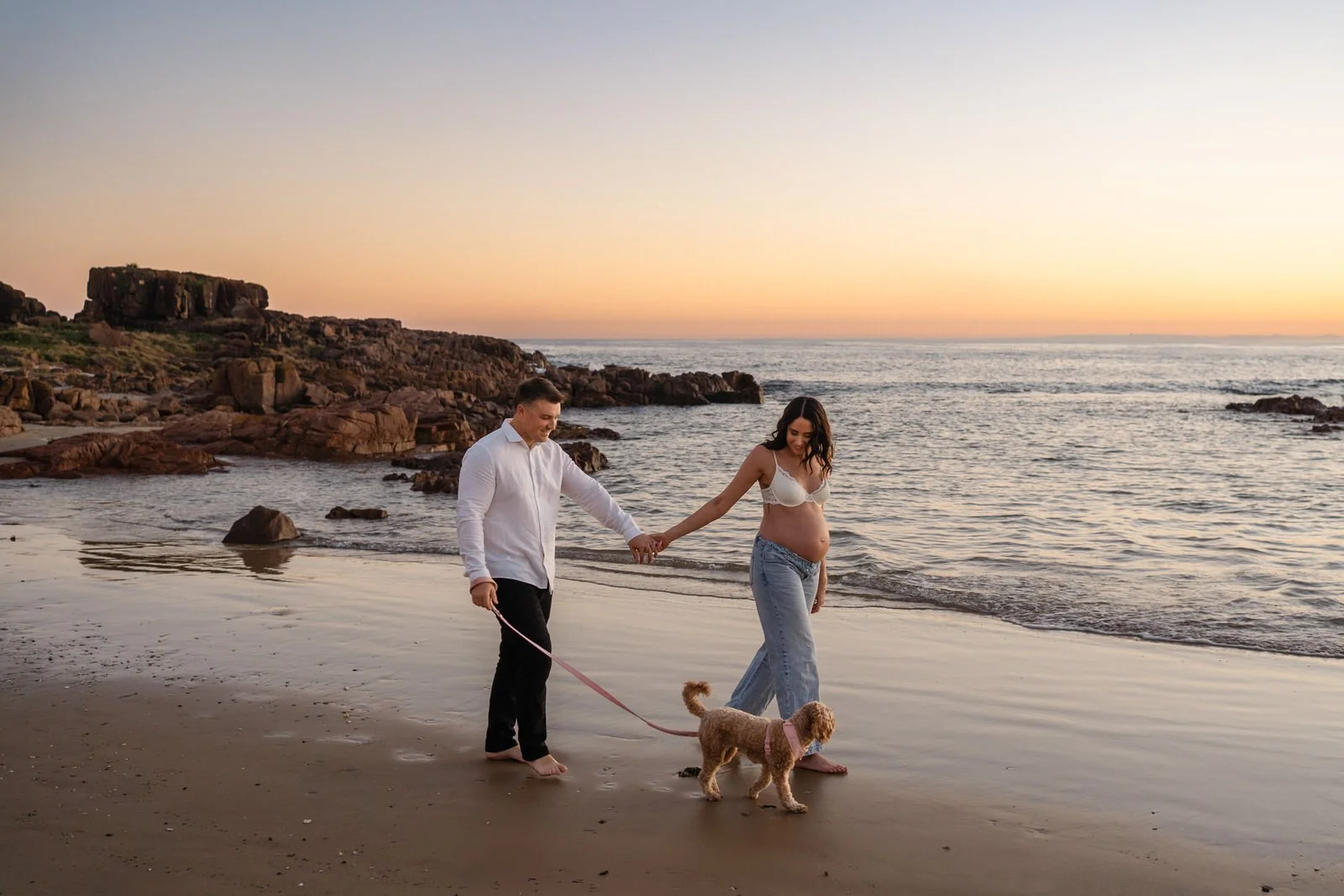A pregnant woman and a man walking their dog at the beach during sunset.