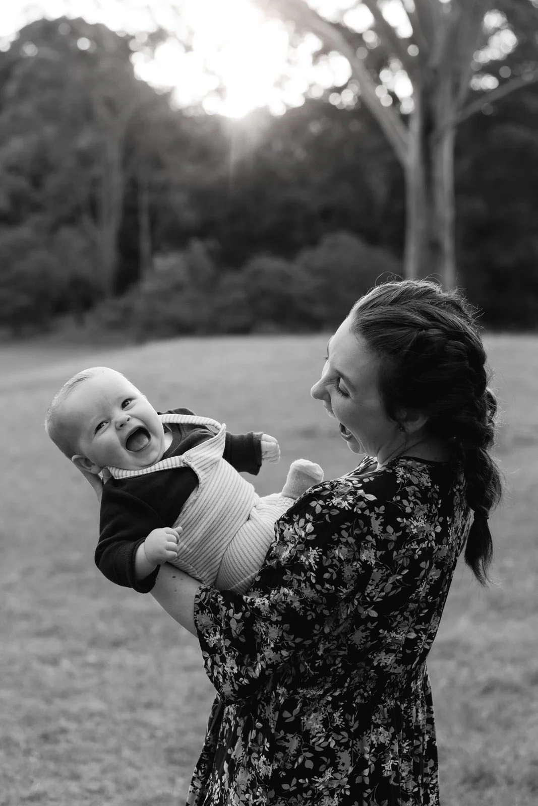 A woman holding a smiling baby outdoors in a grassy area with trees in the background.