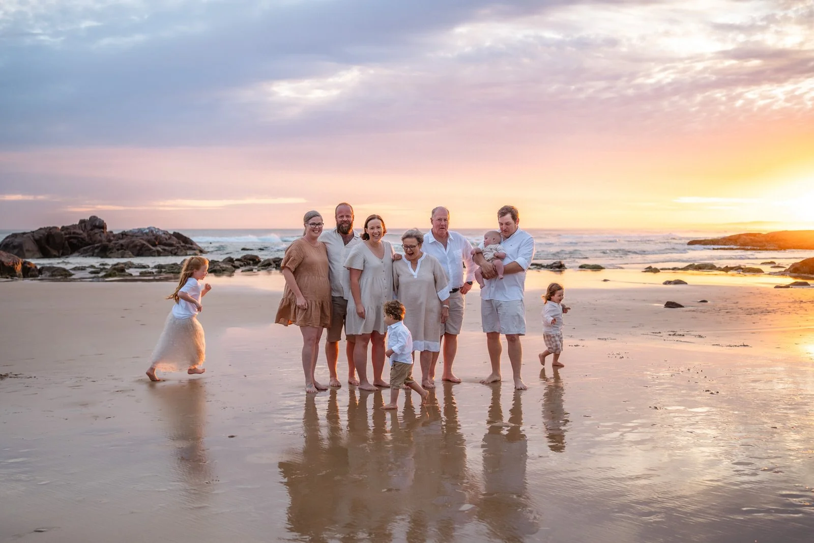 Family of nine standing on the beach during sunset with a rocky shoreline in the background, some children playing and running in the sand.