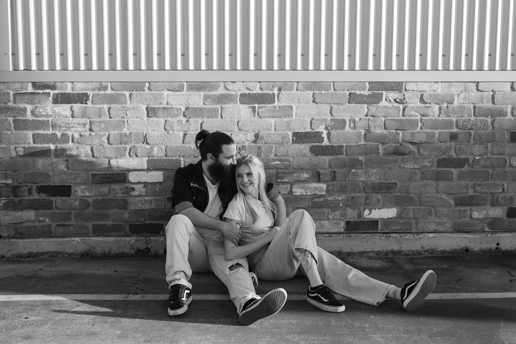 A black and white photo of a smiling young woman and man sitting on the ground against a brick wall. The man has a beard and long hair tied back, wearing a dark jacket and light pants. The woman has long blonde hair, wearing a t-shirt and ripped pant